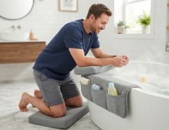 A man using a gray padded bath kneeler and elbow rest set at a bathtub.
